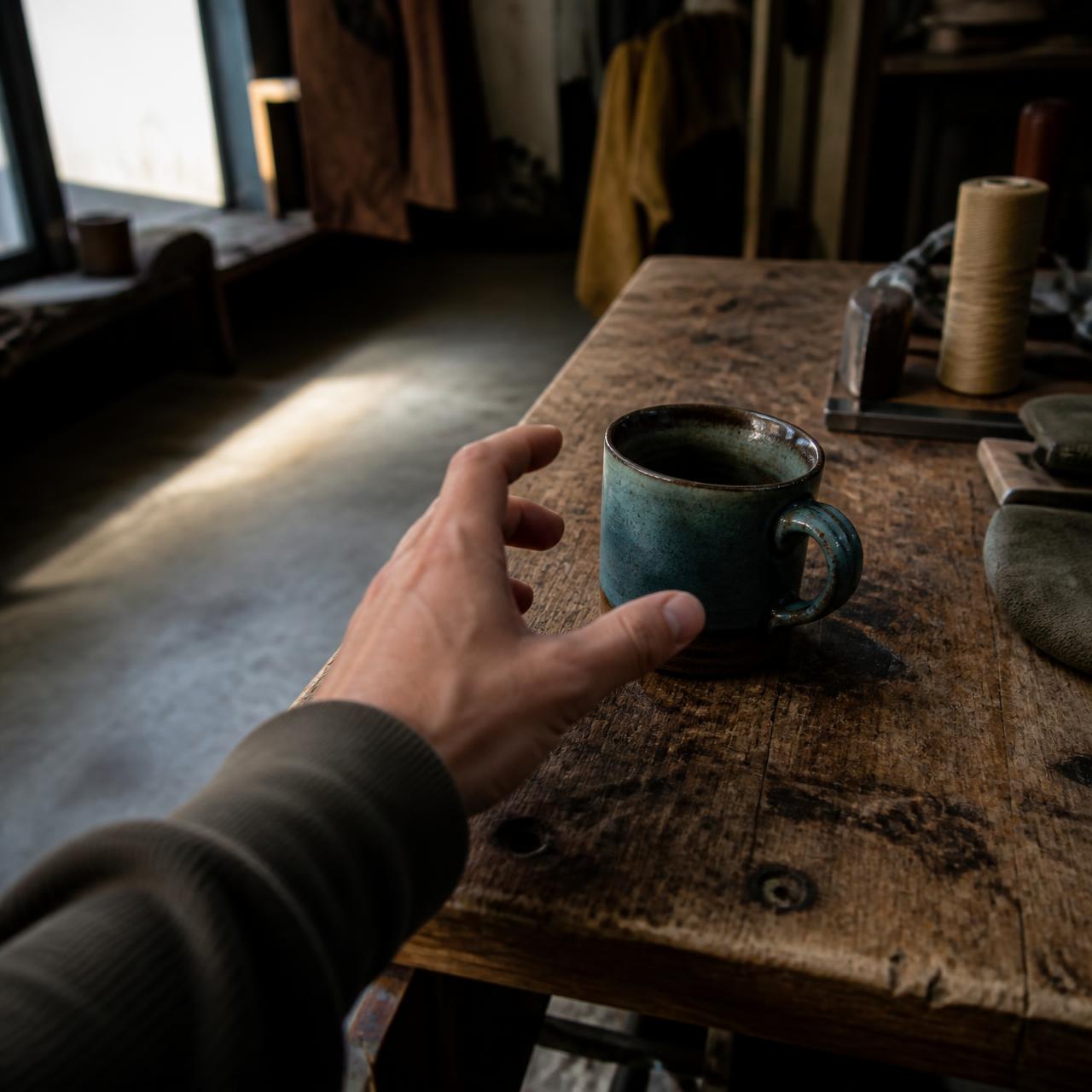 First-person view of a hand reaching for a ceramic mug on a wooden workbench.